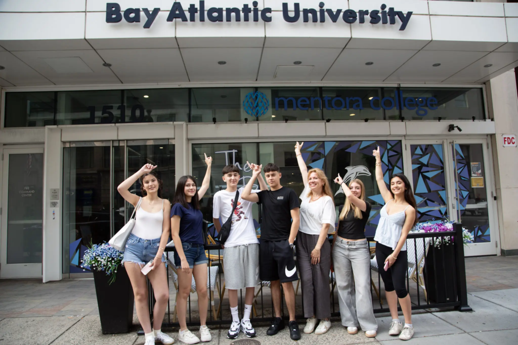 Group of students posing in front of Bay Atlantic University entrance, pointing upwards and smiling.