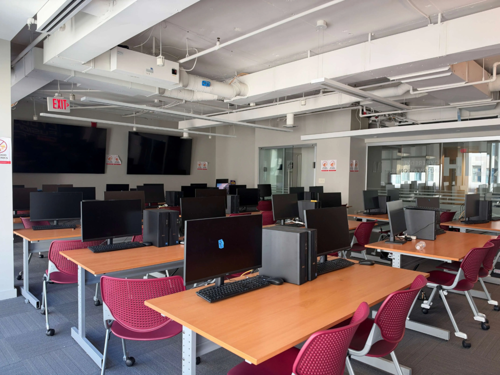 Untitled design (3) Modern computer lab with desktop computers, red chairs, and wooden desks in a bright, spacious room.